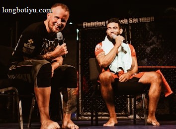 Two individuals are seated on a stage, each holding a microphone and smiling. They are in a sports-related setting with one wearing a casual black outfit and the other in sportswear, with a towel around his neck. The background includes elements of a fighting or martial arts cage.