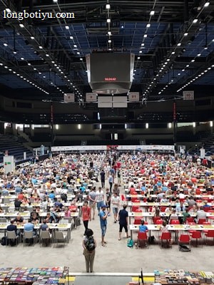 An indoor sports arena filled with rows of tables, each occupied by people engaged in competitive activities. The ceiling is high, with numerous lights illuminating the large space. The audience seating on either side is empty, and there are flags hanging from the upper level.