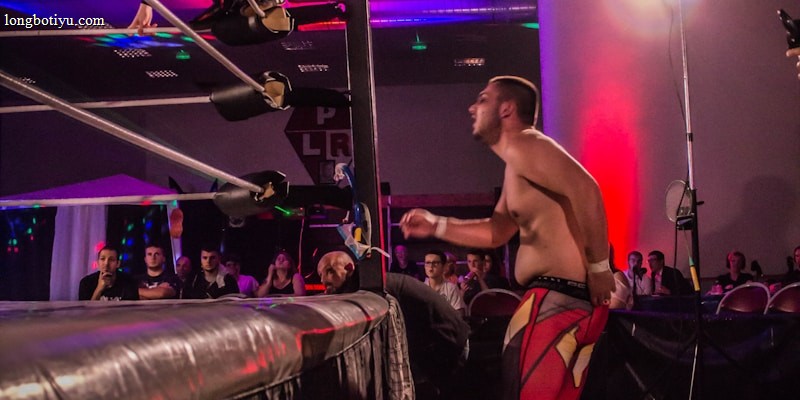 A wrestling match taking place in an indoor venue, where one participant is crouched on the top rope of the ring, while the other stands outside the ring looking at them. A crowd watches the event enthusiastically from their seats.