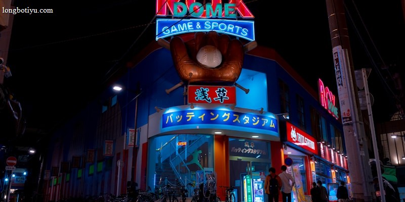 A vibrant, neon-lit facade of a gaming and sports establishment at night, featuring a large glove holding a baseball above the entrance. The building is adorned with colorful signs in both English and Japanese, with various advertisements and an illuminated sign that says 'ROX DOME'. Several people are walking towards the entrance, and bicycles are parked along the side.