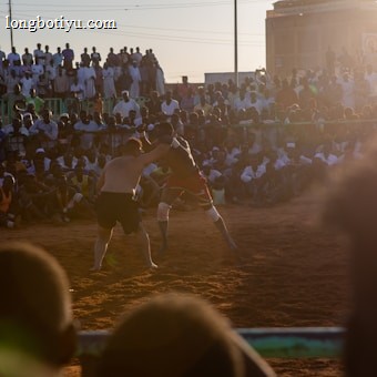 A vibrant wrestling match taking place outdoors, surrounded by a large crowd of spectators. The atmosphere is charged with excitement as people stand and sit around the dirt arena, intently watching the competitors. The sun is setting, casting a warm glow over the scene.